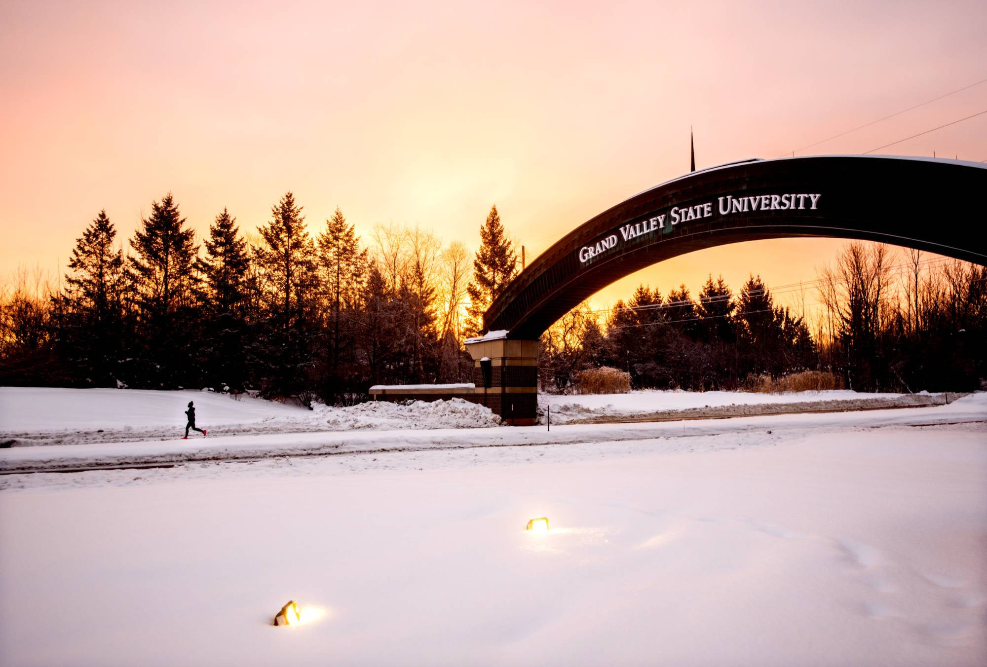 Entrance to GVSU’s Valley Campus in winter, with snow blanketing the surrounding area and a person jogging nearby.